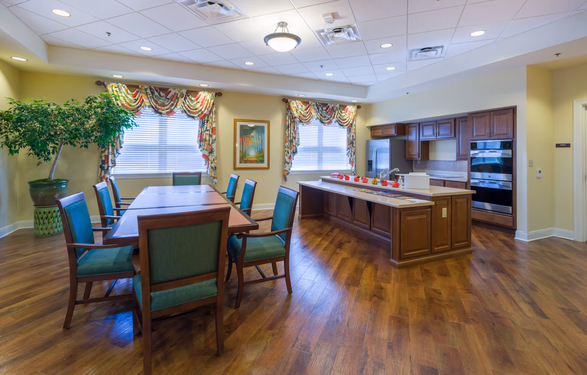 A bright and spacious kitchen and dining area with wooden flooring. The dining area has a rectangular wooden table surrounded by eight green cushioned chairs. The kitchen features a large island with a sink, wooden cabinets, a stainless steel refrigerator, and built-in ovens. Two windows with floral valances allow natural light to fill the room, and a large potted plant is placed near the dining table.