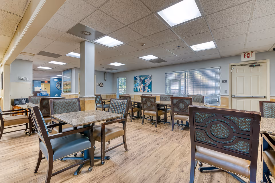 Interior view of a senior living facility dining room with multiple tables and chairs arranged neatly. The room has wood flooring, light-colored walls with a chair rail, and a drop ceiling with fluorescent lighting. There are windows with blinds on one side and an exit door visible in the background. Artwork is hung on the walls, and the space appears clean and well-lit.