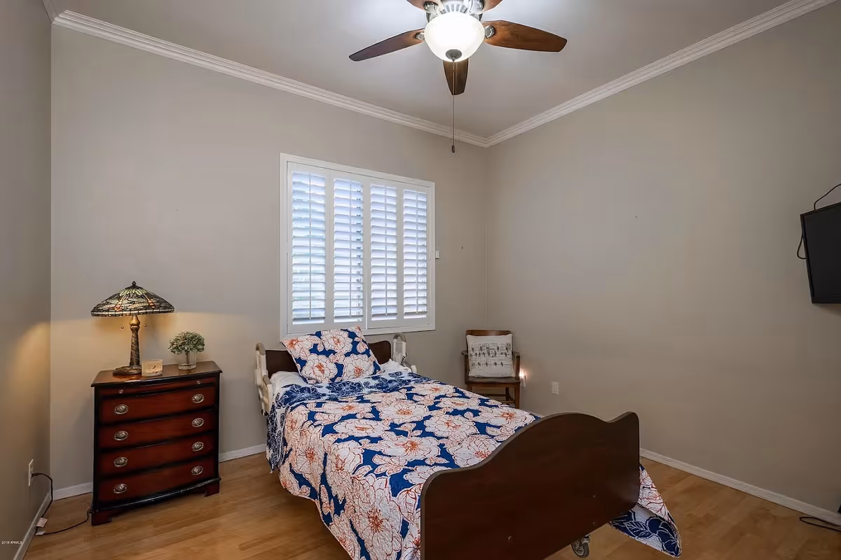 A small bedroom with a single bed covered in a blue and white floral bedspread. There is a wooden nightstand with a decorative lamp and a small plant on it. A wooden chair with a cushion is placed in the corner near a window with white plantation shutters. A ceiling fan with a light is mounted on the ceiling, and a flat-screen TV is mounted on the wall to the right.