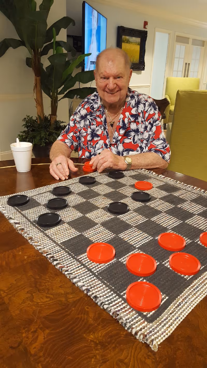 An elderly man wearing a red and white floral shirt is sitting at a wooden table playing checkers with red and black pieces. There is a white cup with a straw on the table, a large green plant in the background, a TV mounted on the wall, and green chairs in the room.