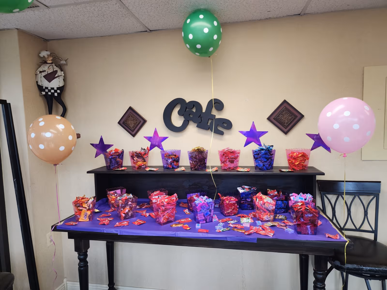 A decorated table with various containers filled with candy and scattered candy pieces on a purple tablecloth. The table is set against a beige wall with the word 'Cafe' in black decorative letters and purple star decorations. Three polka dot balloons in green, pink, and beige are tied to the table and nearby chair. A whimsical chef figurine is mounted on the wall to the left.
