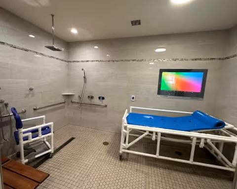 A spacious accessible shower room with beige tiled walls and floor. The room features a white shower chair with blue padding, a blue padded shower bed on a white frame, a handheld showerhead, grab bars, and a wall-mounted TV screen. The lighting is recessed in the ceiling.