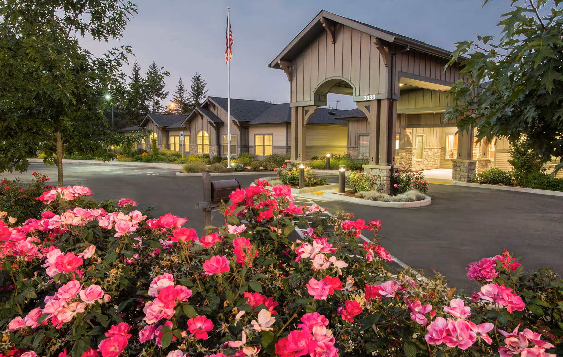 Exterior view of Windsong at Eola Hills facility at dusk, showing the entrance with a covered driveway, illuminated windows, an American flag on a flagpole, and vibrant pink flowers in the foreground.