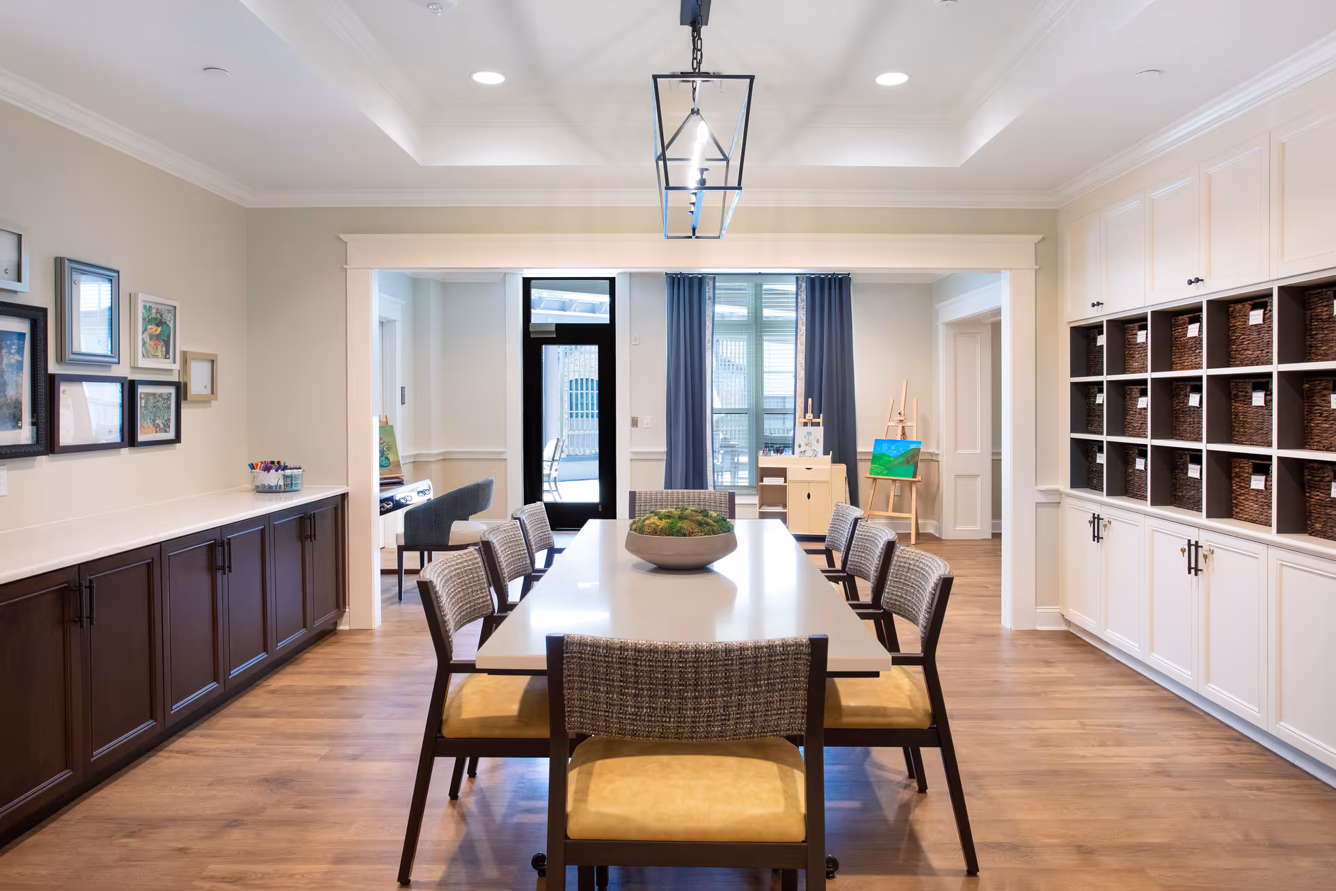 Bright communal dining/activity room with a long table surrounded by chairs, built-in cabinets with baskets, and art supplies by the windows.