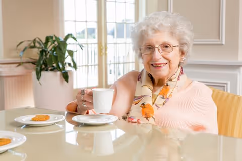 An elderly woman with white hair and glasses sitting at a dining table holding a white cup and smiling. There are plates with cookies on the table and a large window with natural light in the background.