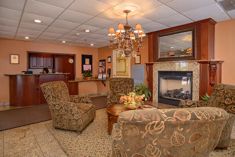 A cozy senior living facility lobby with four patterned armchairs arranged around a round wooden coffee table with a floral centerpiece. There is a fireplace with a large mirror above it on the right side. A chandelier with multiple lampshades hangs from the ceiling. In the background, a reception desk with a staff member is visible, along with framed certificates and a clock on the wall.
