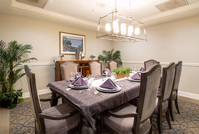 A dining room with a rectangular table covered with a patterned tablecloth and set with plates, glasses, and purple folded napkins. Eight upholstered chairs surround the table. A modern chandelier hangs above, and there are plants and a framed picture on the wall in the background.