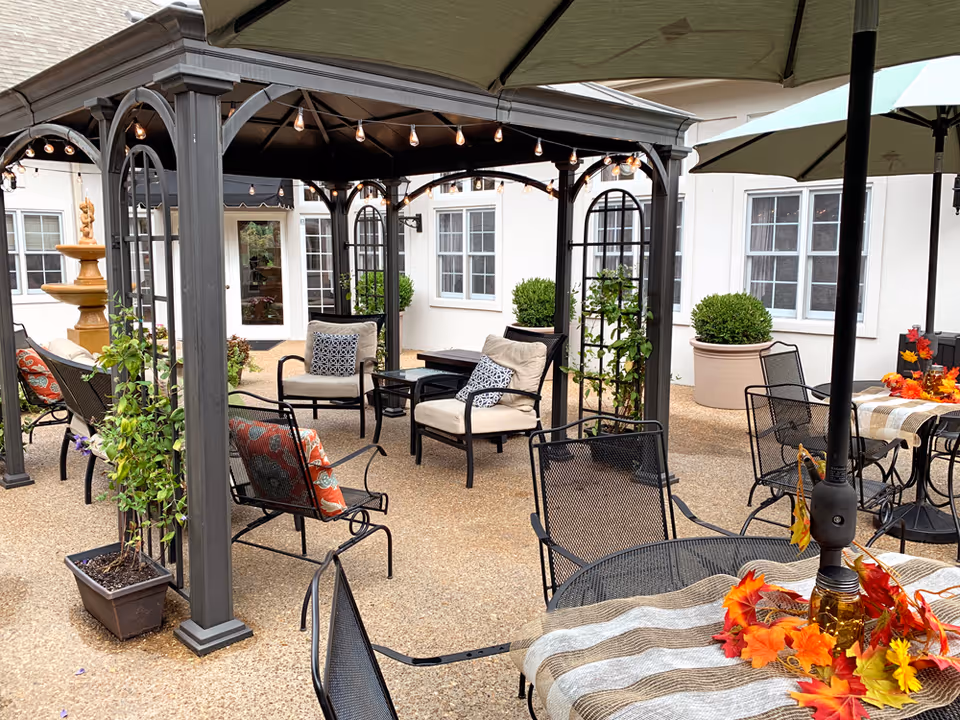 Outdoor patio area with a gazebo featuring string lights, cushioned chairs, metal chairs and tables with umbrellas, potted plants, and a decorative fountain in the background.