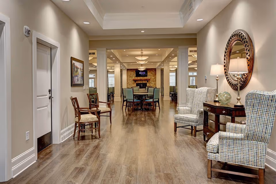A spacious, well-lit interior hallway in a senior living facility with wooden flooring, beige walls, and white trim. On the right side, there are two patterned armchairs with a wooden side table between them, topped with a lamp and decorative items, and a round mirror above. On the left side, there are two wooden chairs and a framed painting on the wall. In the background, there is a dining area with tables and chairs, and a stone fireplace with a TV mounted above it.