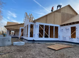 A partially constructed building wrapped in Tyvek with construction materials on the ground and workers on the roof under a blue sky.