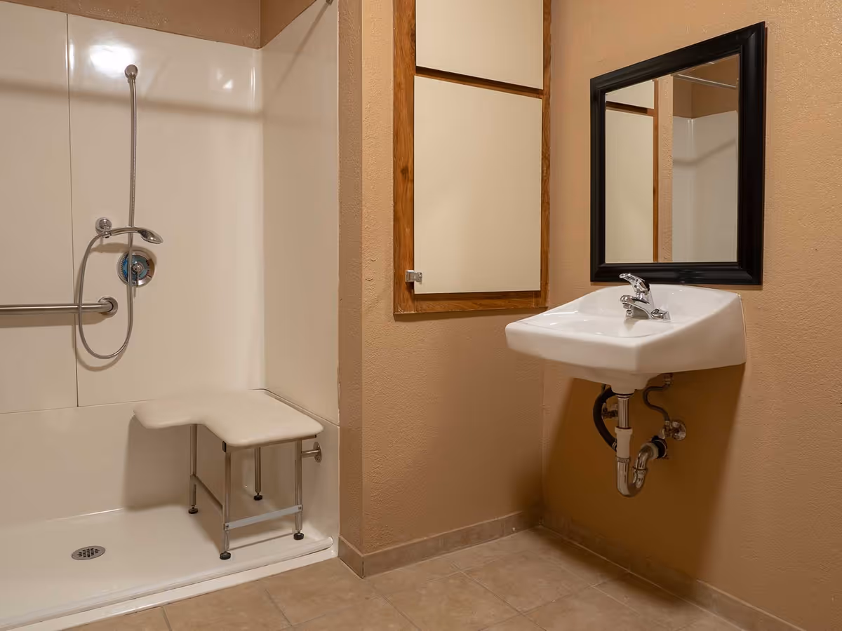 A bathroom with a walk-in shower featuring a handheld showerhead and a built-in bench. There is a wall-mounted sink with exposed plumbing underneath and a rectangular mirror above it. The walls are painted beige and the floor is tiled.