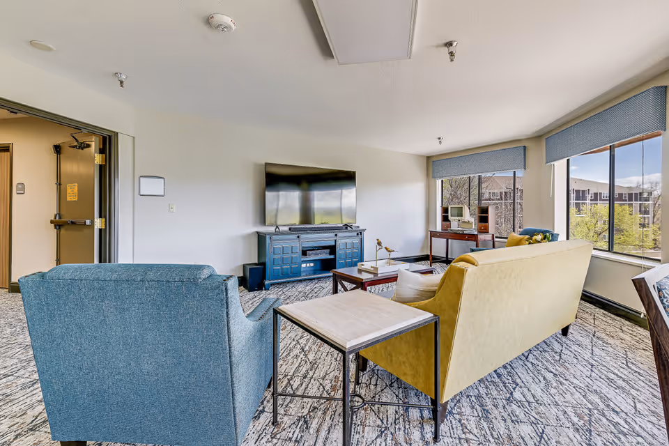 A bright living room with large windows showing an outdoor view. The room features a blue armchair, a yellow sofa, a wooden coffee table, and a blue TV stand with a flat-screen television mounted on the wall. There is also a small wooden desk with a vintage computer near the windows.