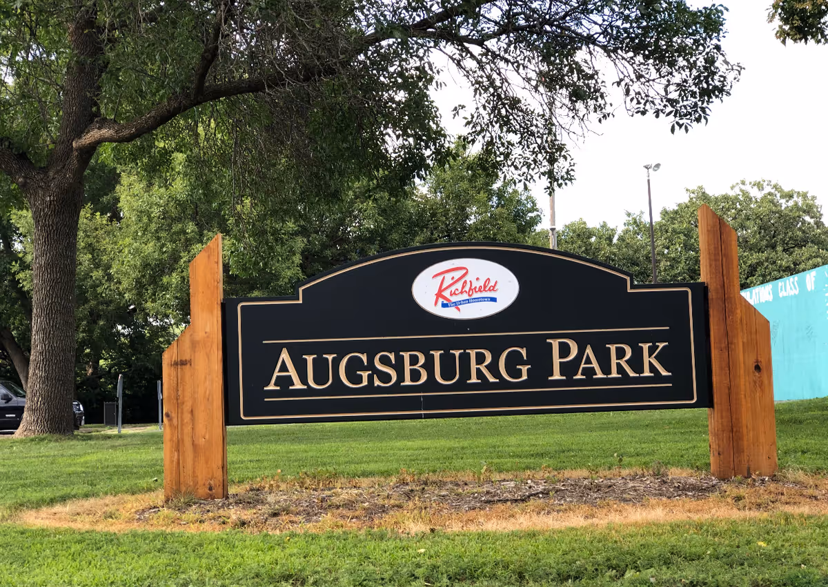 A large black and gold sign reading 'Augsburg Park' mounted on two wooden posts in a grassy area with trees in the background.