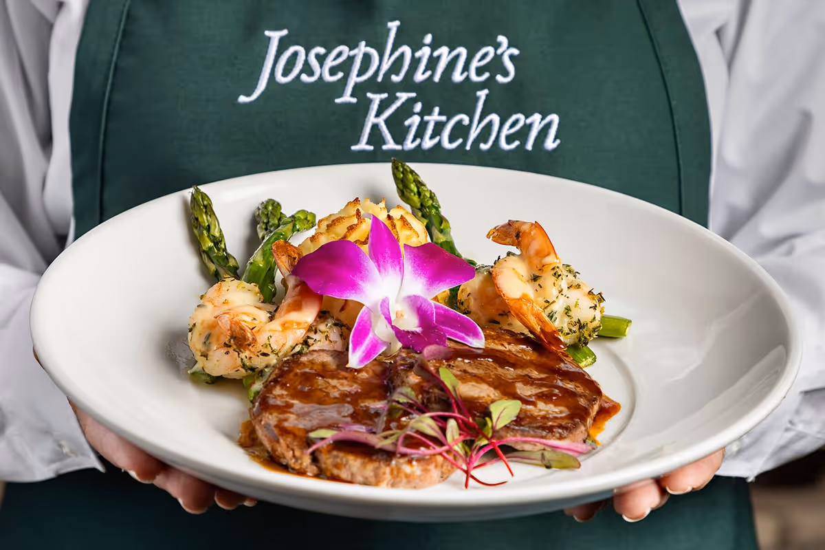 A person wearing a green apron with the text 'Josephine's Kitchen' holds a white plate with a gourmet meal consisting of grilled shrimp, asparagus, mashed potatoes, a meat patty with sauce, and a purple edible flower garnish.