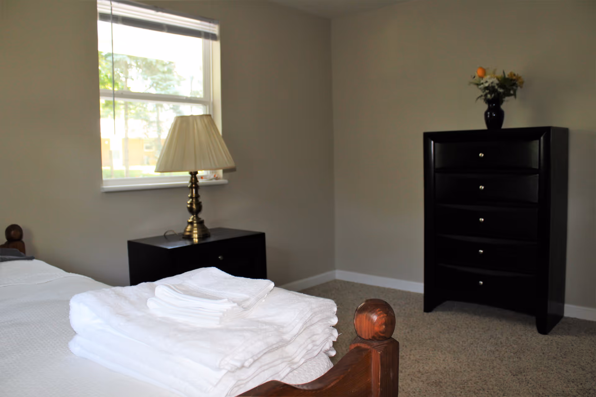 A bedroom with a wooden bed frame, a neatly folded stack of white towels on the bed, a black nightstand with a brass lamp, a window with blinds partially open, and a black chest of drawers with a vase of flowers on top.