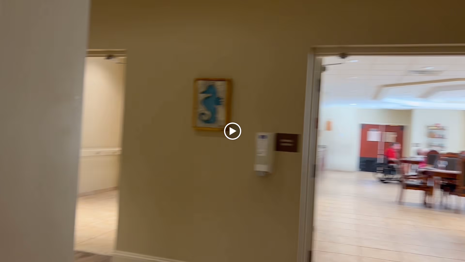 View through a hallway into a dining area with tables and chairs. A person in a red shirt is seated at one of the tables. The walls are beige, and there is a framed picture of a blue seahorse on the wall near the hallway entrance.