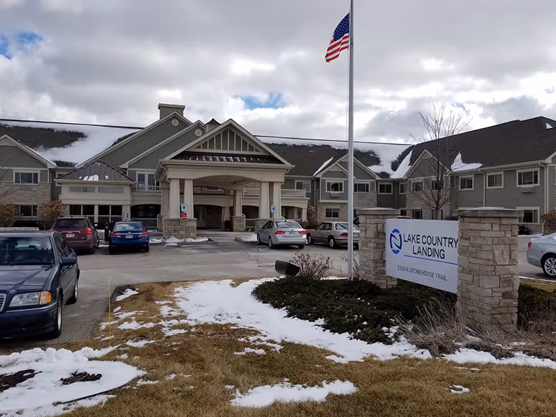 Front exterior view of Lake Country Landing facility with a parking lot in front, several cars parked, an American flag on a flagpole, and a sign displaying the facility name and address. Snow patches are visible on the ground and roof under a cloudy sky.