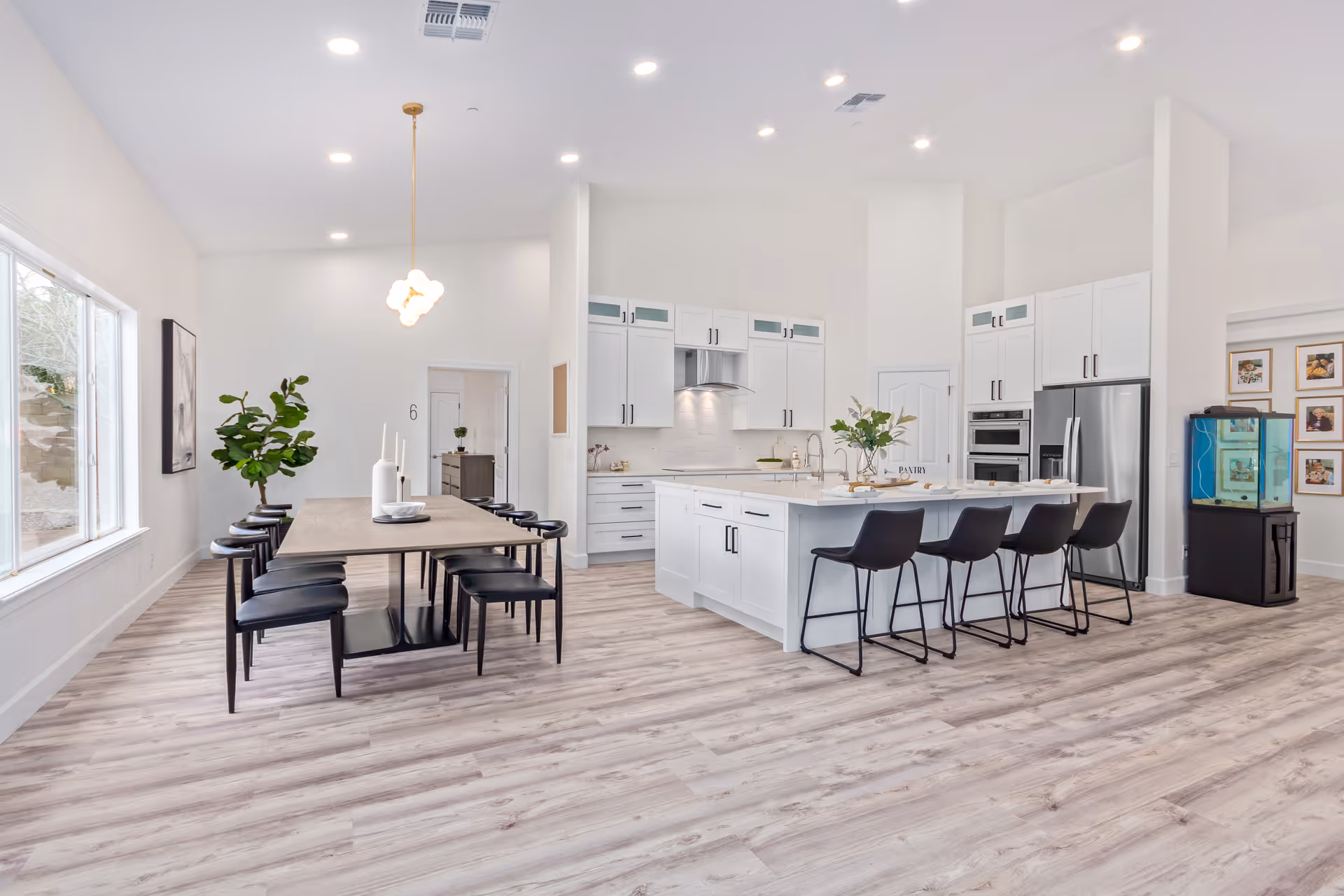 Bright and modern open-concept kitchen and dining area with white cabinetry, a large kitchen island with black bar stools, a wooden dining table with black chairs, a large window, and light wood flooring.