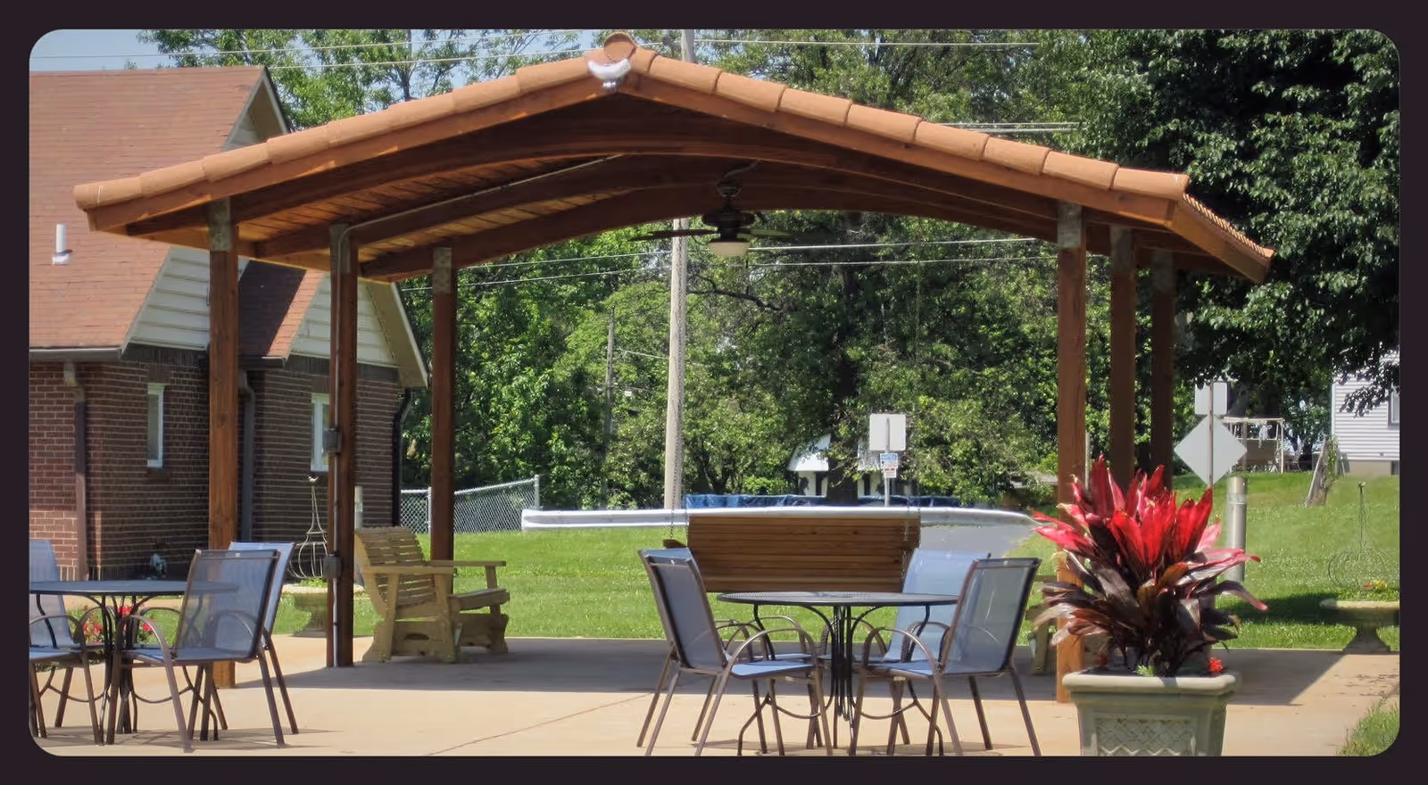 Outdoor covered patio area with a wooden roof supported by wooden posts, featuring a round table with four chairs and a wooden bench. There is a large planter with red and green foliage on the right side, and a brick building partially visible on the left. Green grass and trees are in the background under a clear sky.