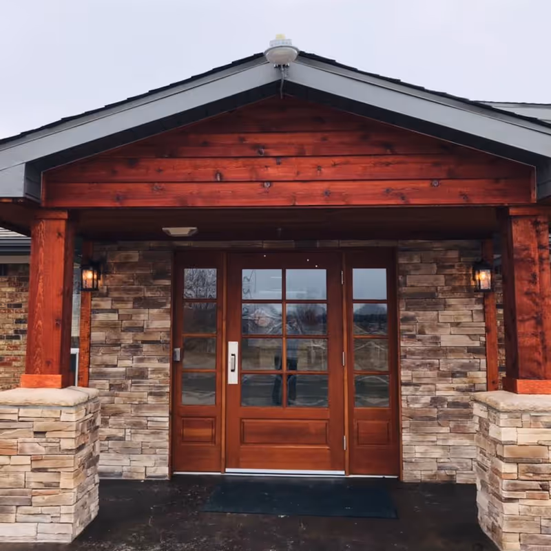 Front entrance of a building with wooden double doors featuring glass panes, stone pillars, and a wooden overhang with a light fixture on each side.