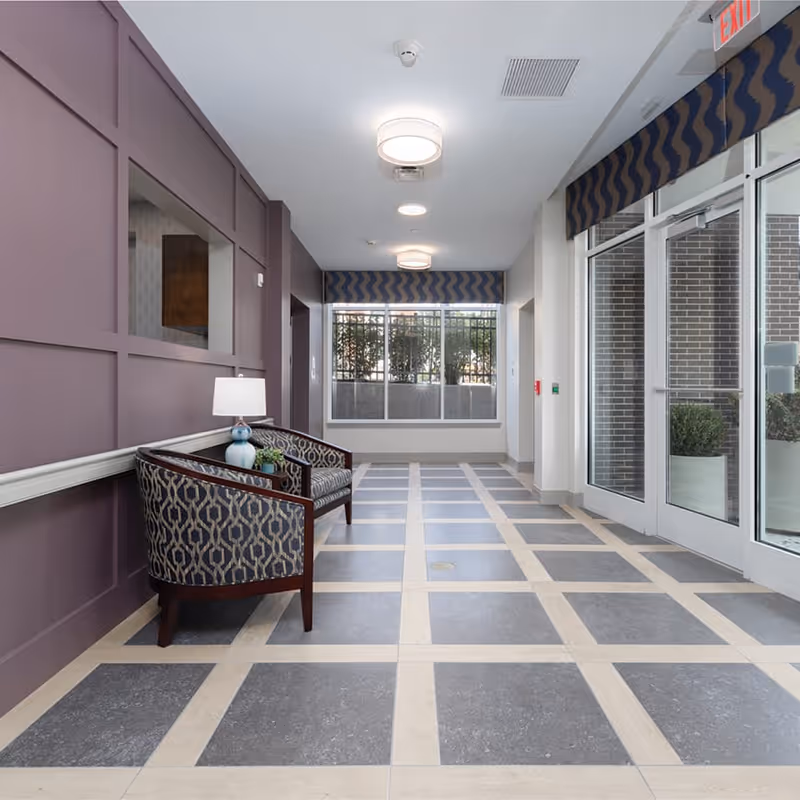 A bright and clean hallway in a senior living facility with patterned tile flooring and purple paneled walls. Two upholstered chairs with a small table and lamp are placed against the left wall. Large windows and glass doors on the right side allow natural light to enter, with a view of outdoor plants and a brick wall outside. Ceiling lights and an exit sign are visible.