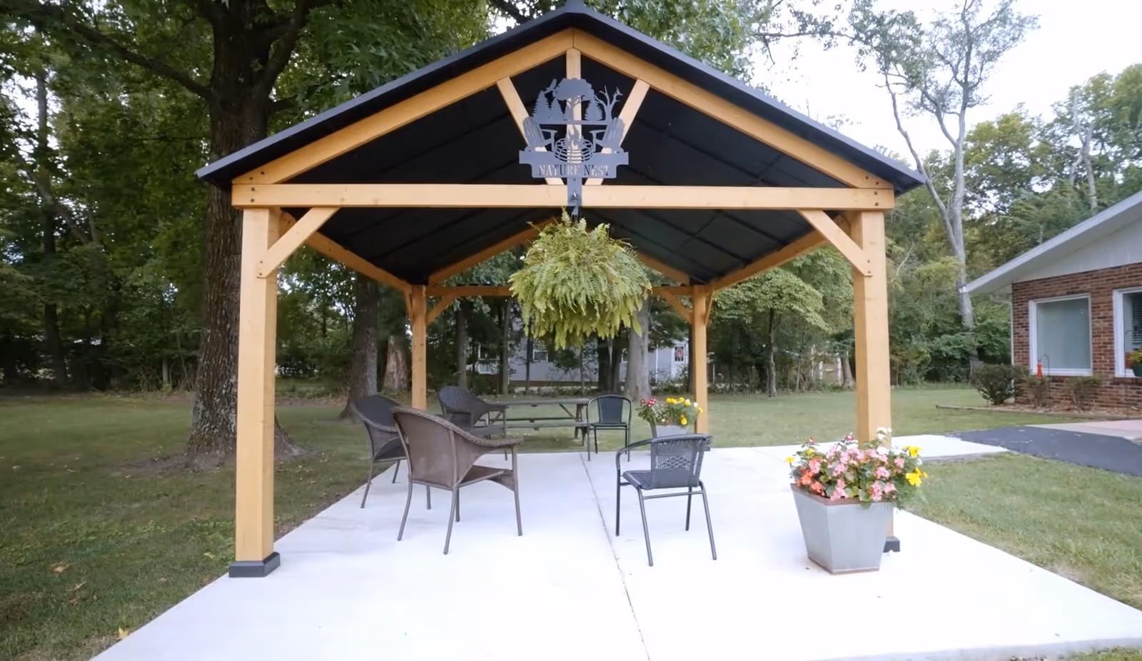 Outdoor wooden pavilion with a black roof, featuring several chairs and a picnic table underneath. A hanging fern plant is suspended from the center beam, and a metal sign with the words 'Nature Nest' and a nature-themed design is mounted at the peak of the pavilion. The pavilion is situated on a concrete slab surrounded by grass and trees, with a building visible to the right.