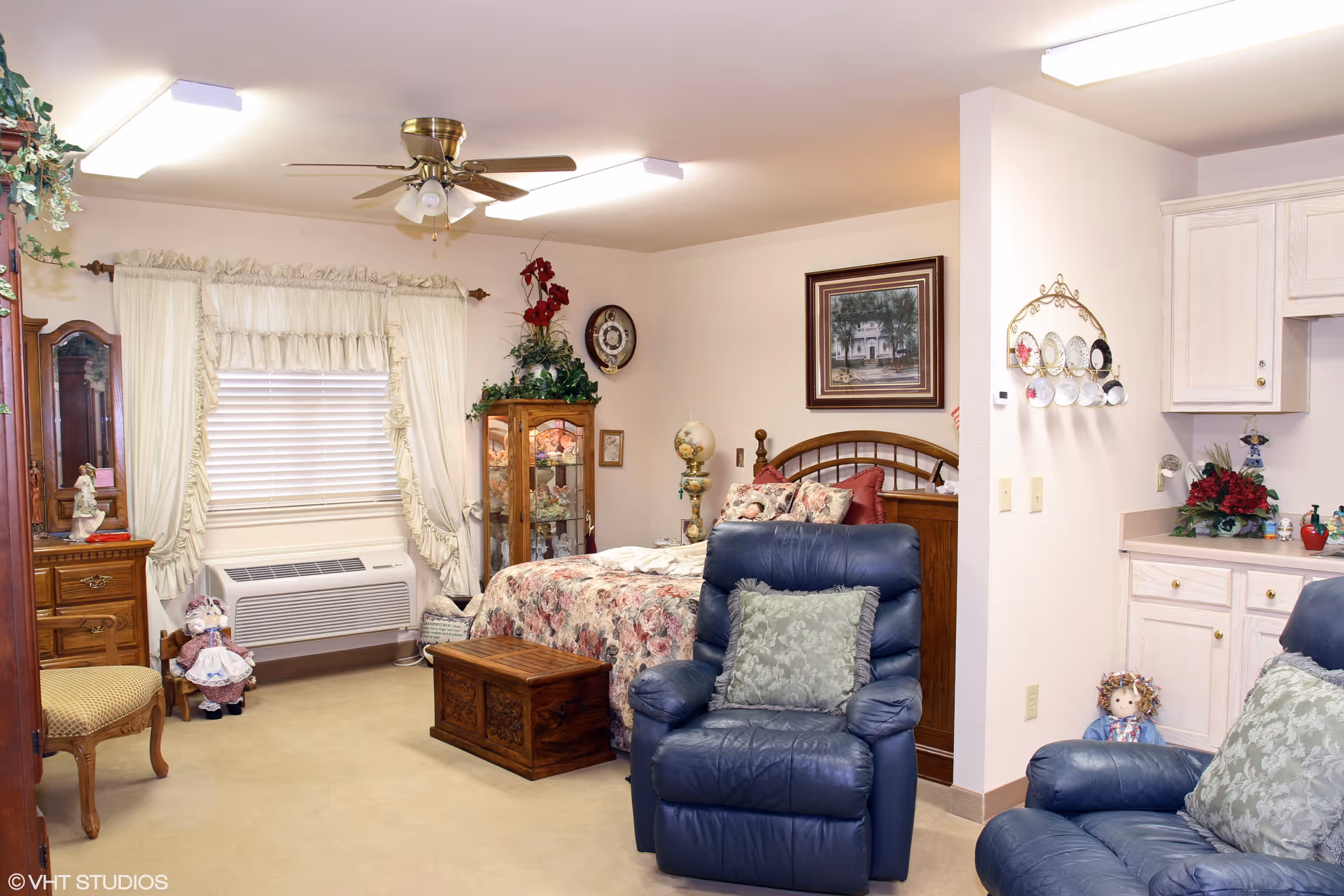 A cozy bedroom in an assisted living facility featuring a bed with floral bedding, a wooden chest at the foot of the bed, a blue recliner chair with a green pillow, a wooden dresser with a mirror, and a window with white curtains. The room also has a ceiling fan, decorative plants, and a small kitchenette area with white cabinets and a countertop.
