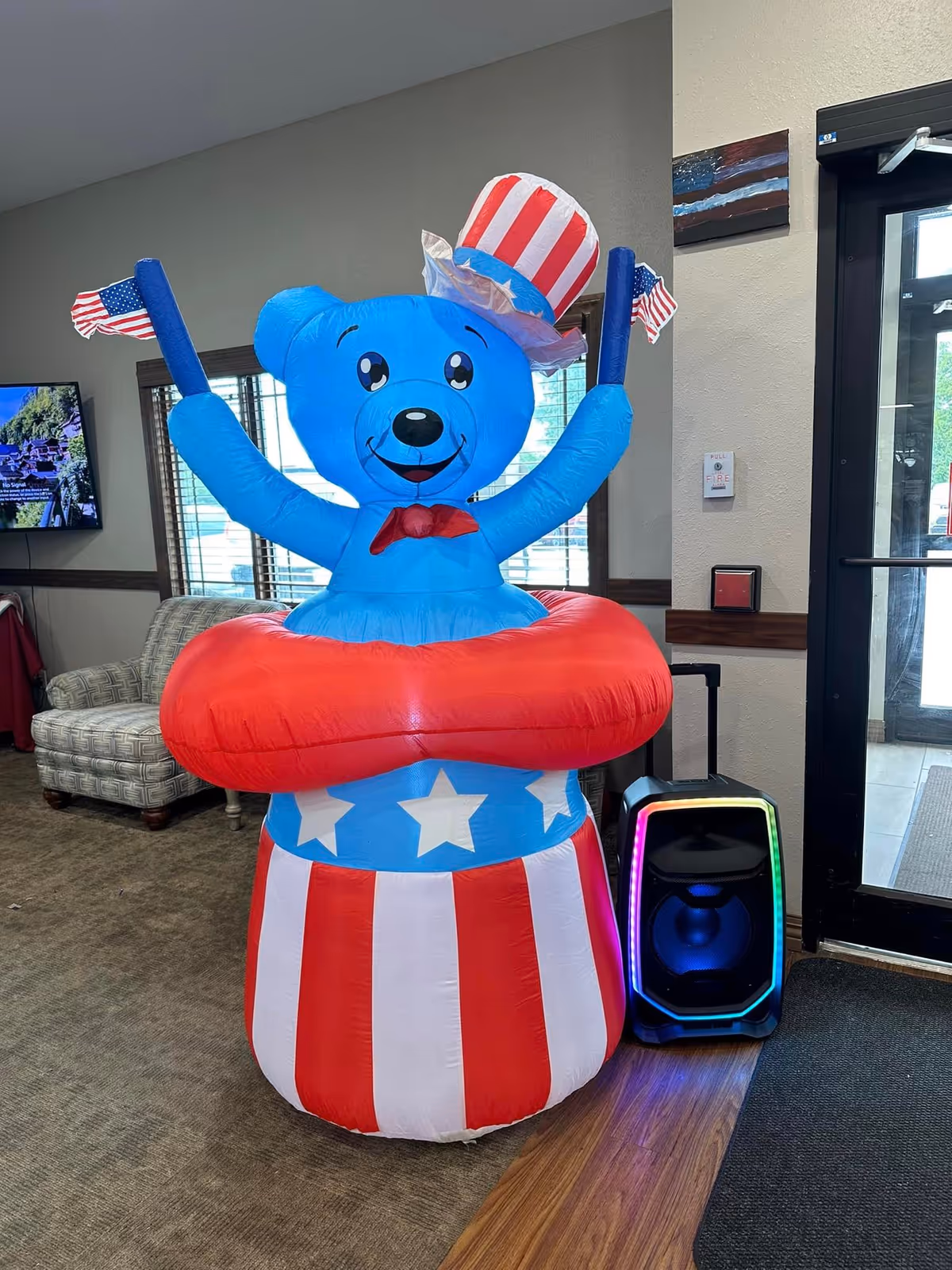 An inflatable blue bear wearing a red, white, and blue Uncle Sam hat and bow tie, holding two small American flags, standing inside a red and white striped float with white stars. The bear is placed in a room with a patterned armchair, a TV mounted on the wall, a door with glass panels, and a speaker with colorful LED lights on the floor next to it.