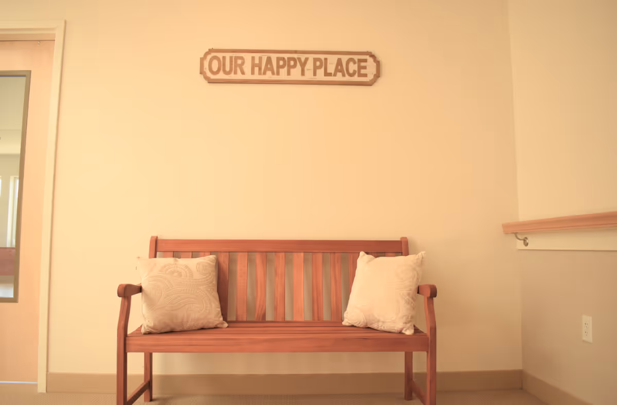 A wooden bench with two beige patterned cushions placed against a light-colored wall. Above the bench is a wooden sign that reads 'OUR HAPPY PLACE'. To the left is a wooden door with a glass panel, and to the right is a handrail attached to the wall.
