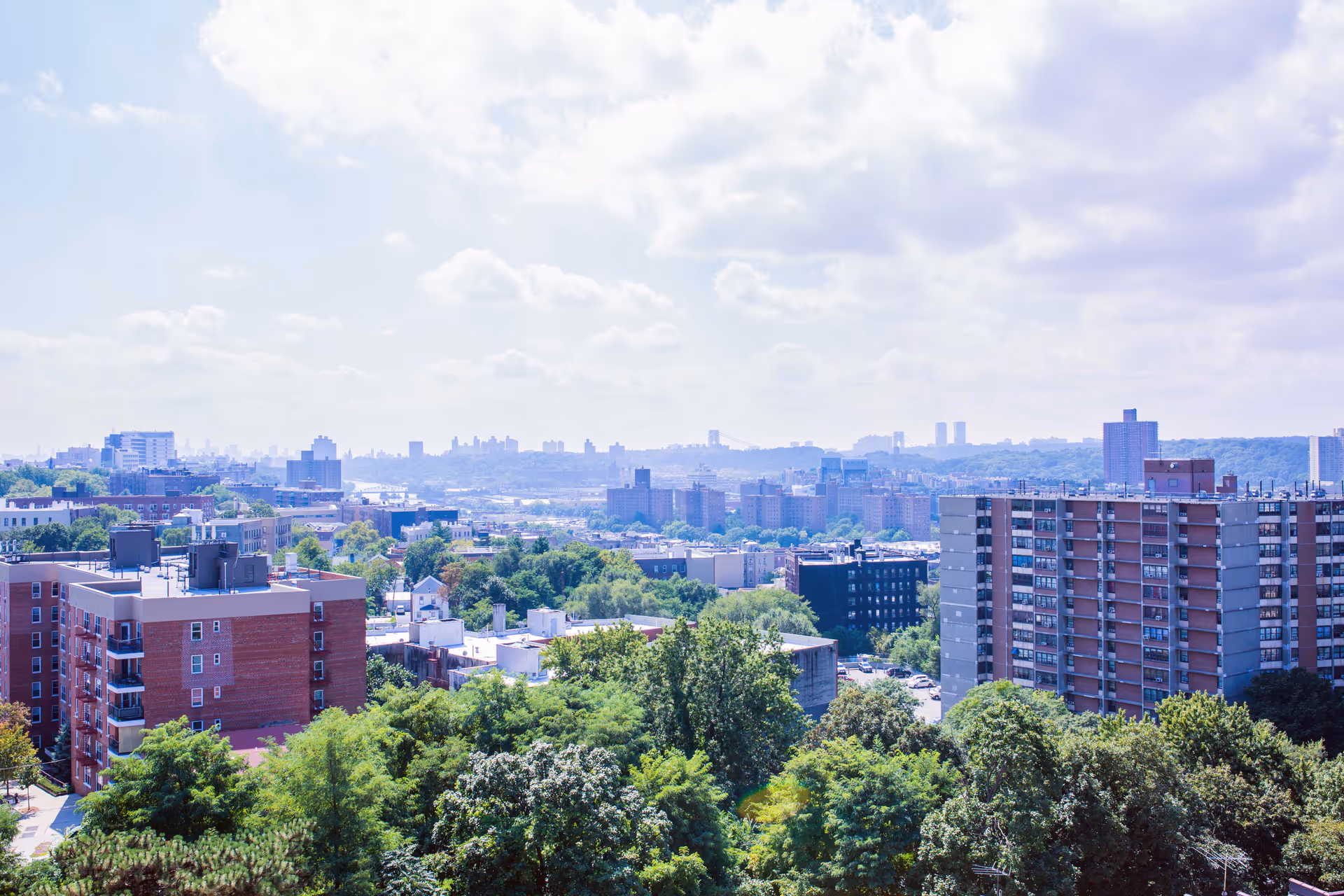 A cityscape view showing multiple residential buildings and apartment complexes surrounded by green trees under a partly cloudy sky.