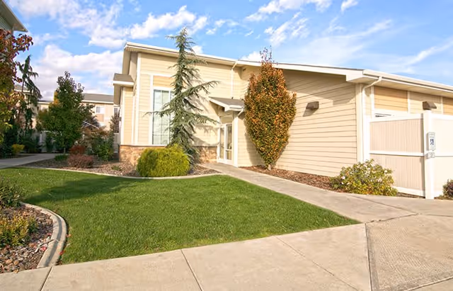 Exterior view of a single-story building with beige siding and a small landscaped lawn with bushes and trees under a partly cloudy sky.