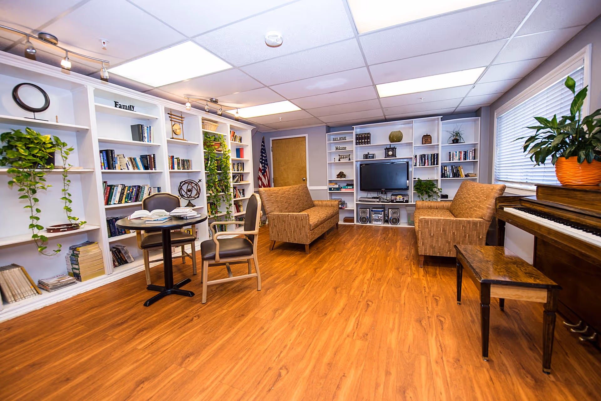 A cozy living room area in Uptown Care Center featuring wooden flooring, two brown cushioned armchairs, a wooden piano with a bench, and a round table with two chairs. The walls are lined with white built-in bookshelves filled with books, plants, and decorative items. An American flag stands near a closed door, and a flat-screen TV is mounted on the shelves. Large windows with blinds allow natural light to brighten the room.