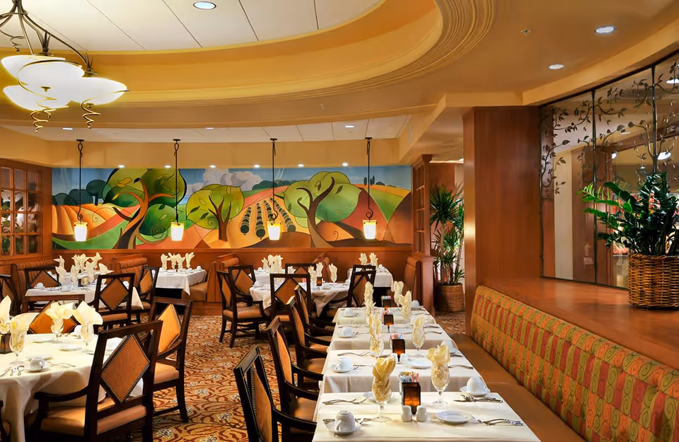 A warmly lit dining room with neatly arranged tables covered in white tablecloths, each set with folded napkins, cups, and glasses. The room features wooden chairs with cushioned seats, a patterned carpet, and a colorful mural of trees and fields on the far wall. Pendant lights hang from the ceiling, and there are potted plants near a wooden partition with decorative glass panels.