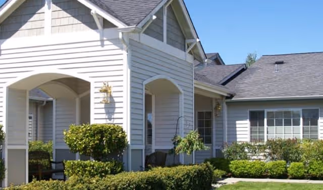 Exterior view of a single-story building with white siding and a gray shingled roof, featuring a covered porch area with benches and surrounded by neatly trimmed bushes and greenery under a clear blue sky.