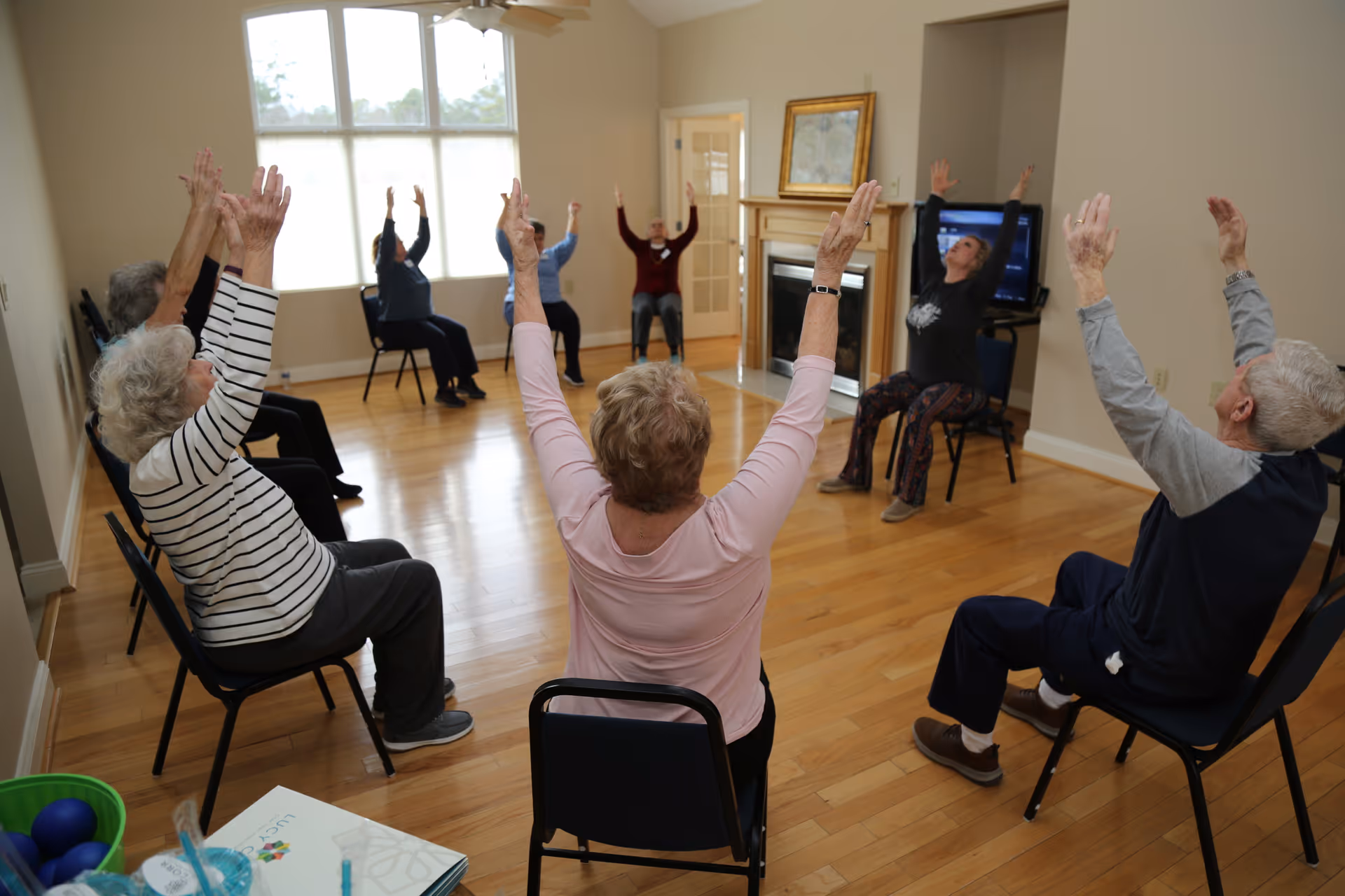 A group of elderly people seated in a circle on chairs in a bright room with wooden floors, participating in a seated exercise or stretching activity with their arms raised. The room has large windows, a fireplace, and a television.