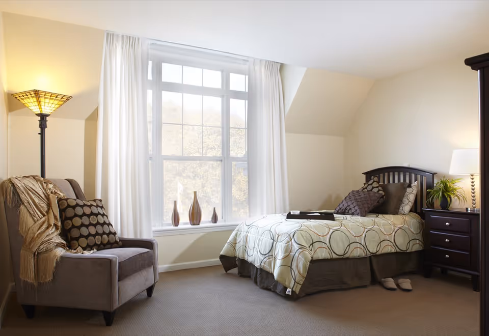 A cozy bedroom with a single bed covered in a patterned comforter and multiple pillows. Next to the bed is a dark wooden nightstand with a lamp and a small plant. A comfortable armchair with a throw blanket and a decorative pillow is positioned near a large window with white curtains. Three decorative vases are placed on the windowsill. The room has beige walls and carpeted floor.