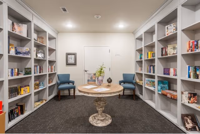A cozy reading room with built-in white bookshelves filled with books and board games on both sides. In the center, there is a round wooden table with a decorative plant and papers on it. Two blue upholstered chairs are placed against the back wall, which has a closed white door and a wall clock.