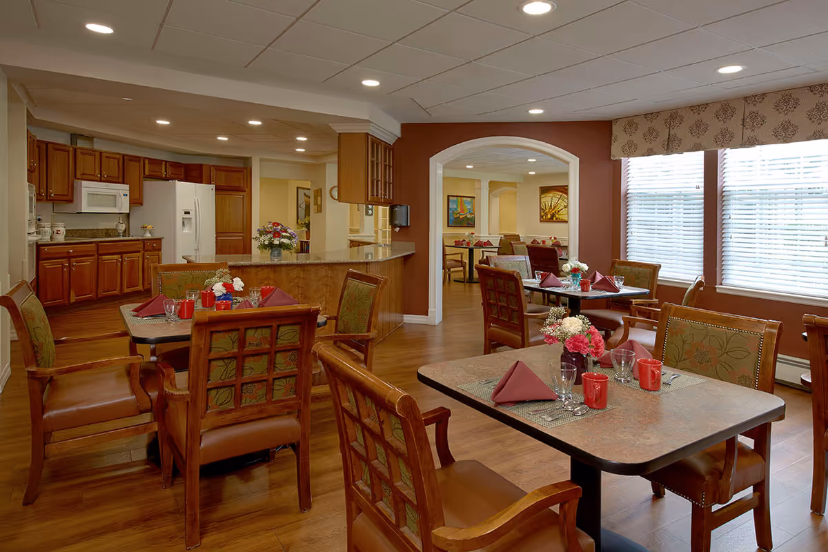 A warm and inviting dining area in a senior living facility featuring several tables set with red napkins, glassware, and red mugs. The room has wooden chairs with cushioned seats, large windows with blinds, and a kitchen area with wooden cabinets, a white refrigerator, and a microwave in the background. The walls are painted in warm tones with decorative artwork and floral arrangements on the tables.