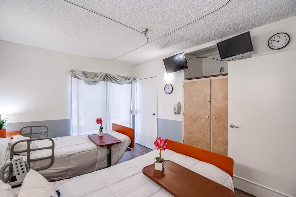 A bright room with two hospital-style beds, each with a brown overbed table holding a small potted pink flower. The beds have white bedding and one has a bed rail and remote control. There is a window with sheer curtains, a closed door, a wall-mounted clock, a hand sanitizer dispenser, a wooden cabinet, and two wall-mounted televisions.