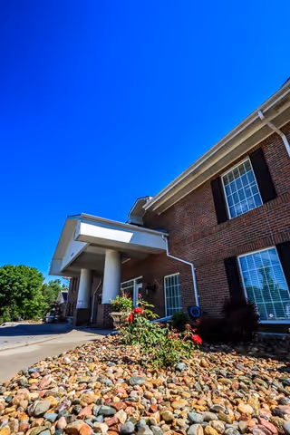 Exterior view of a brick building with white columns at the entrance, surrounded by a landscaped area with rocks and some plants under a clear blue sky.