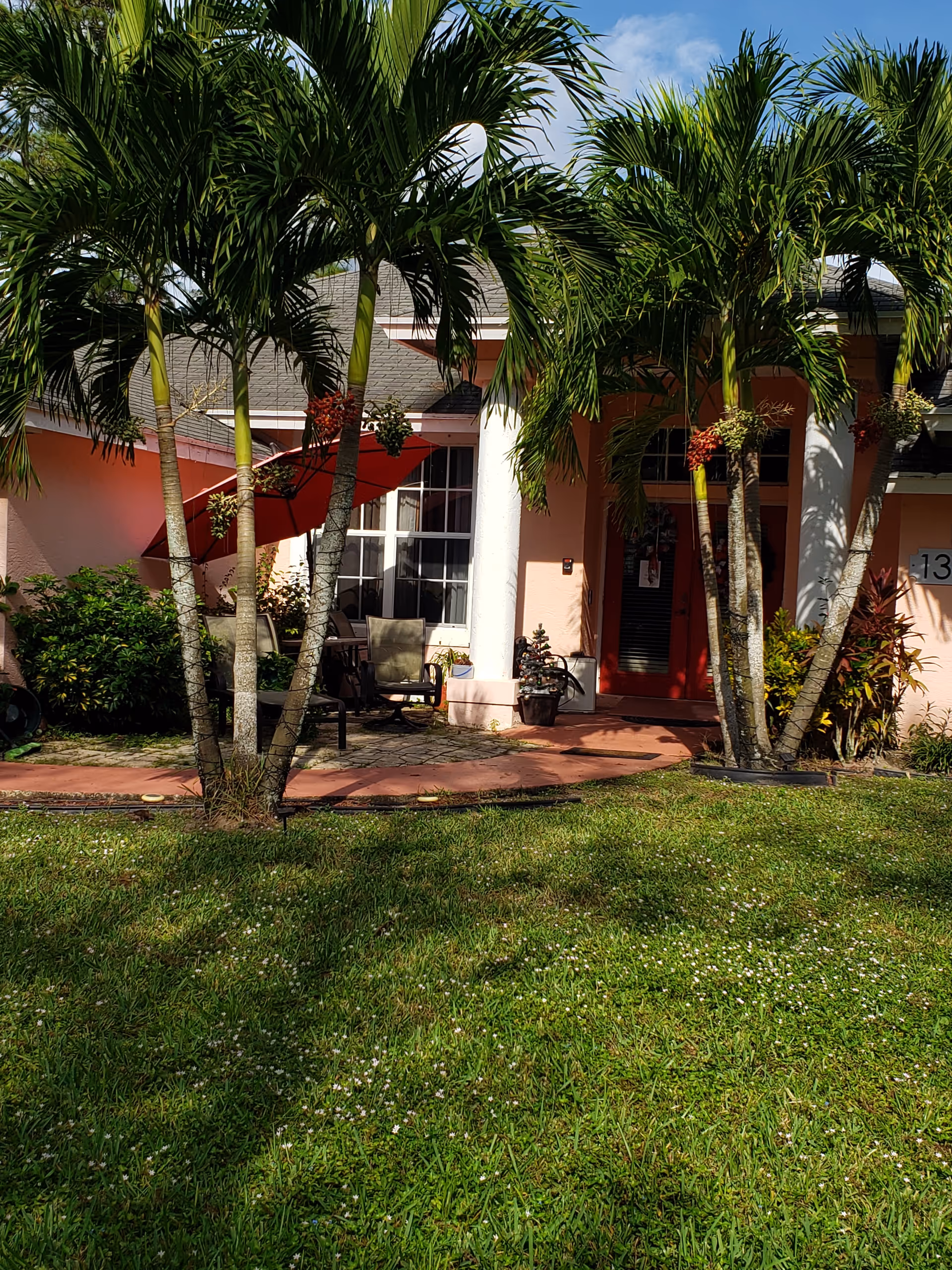 Front entrance of a pink single-story building with palm trees, a grassy lawn, and a small patio with chairs and an umbrella.