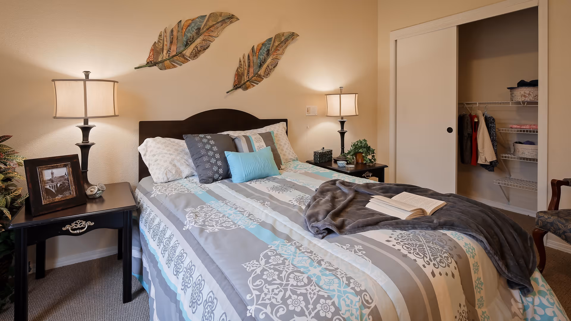 A cozy bedroom featuring a neatly made bed with patterned bedding in shades of gray, white, and blue. There are decorative pillows and a gray throw blanket with an open book on the bed. On either side of the bed are black nightstands with matching lamps. One nightstand has a framed photo and a small clock, while the other has a plant and decorative items. Above the bed, two large decorative feathers hang on the wall. In the background, there is an open closet with clothes and shelves.