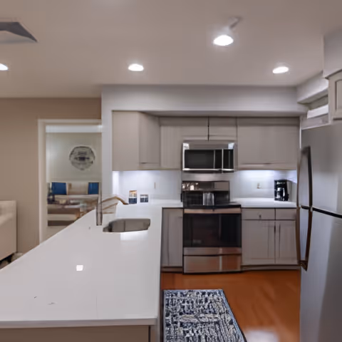 Modern kitchen with white countertops, stainless steel appliances including a microwave, oven, and refrigerator. The kitchen has white cabinets and a blue patterned rug on the wooden floor. Recessed ceiling lights illuminate the space, and a living room area is visible through an open doorway.