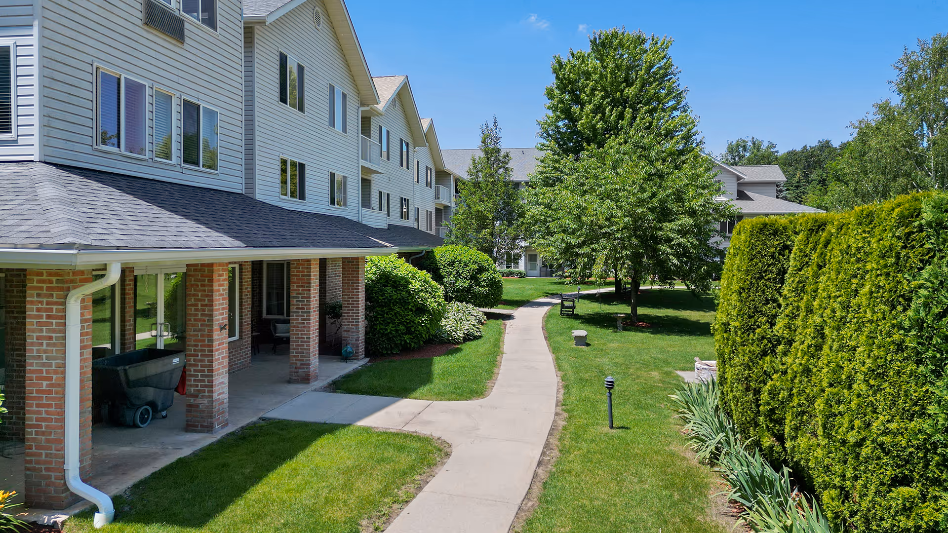 Outdoor view of Holiday Blue Water Lodge showing a paved walkway surrounded by green grass, bushes, and trees. The building has a brick and siding exterior with multiple windows and a covered porch area.