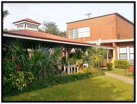 Exterior view of Crestview Manor showing a brick building with large windows and a covered porch area surrounded by green plants and a well-maintained lawn.