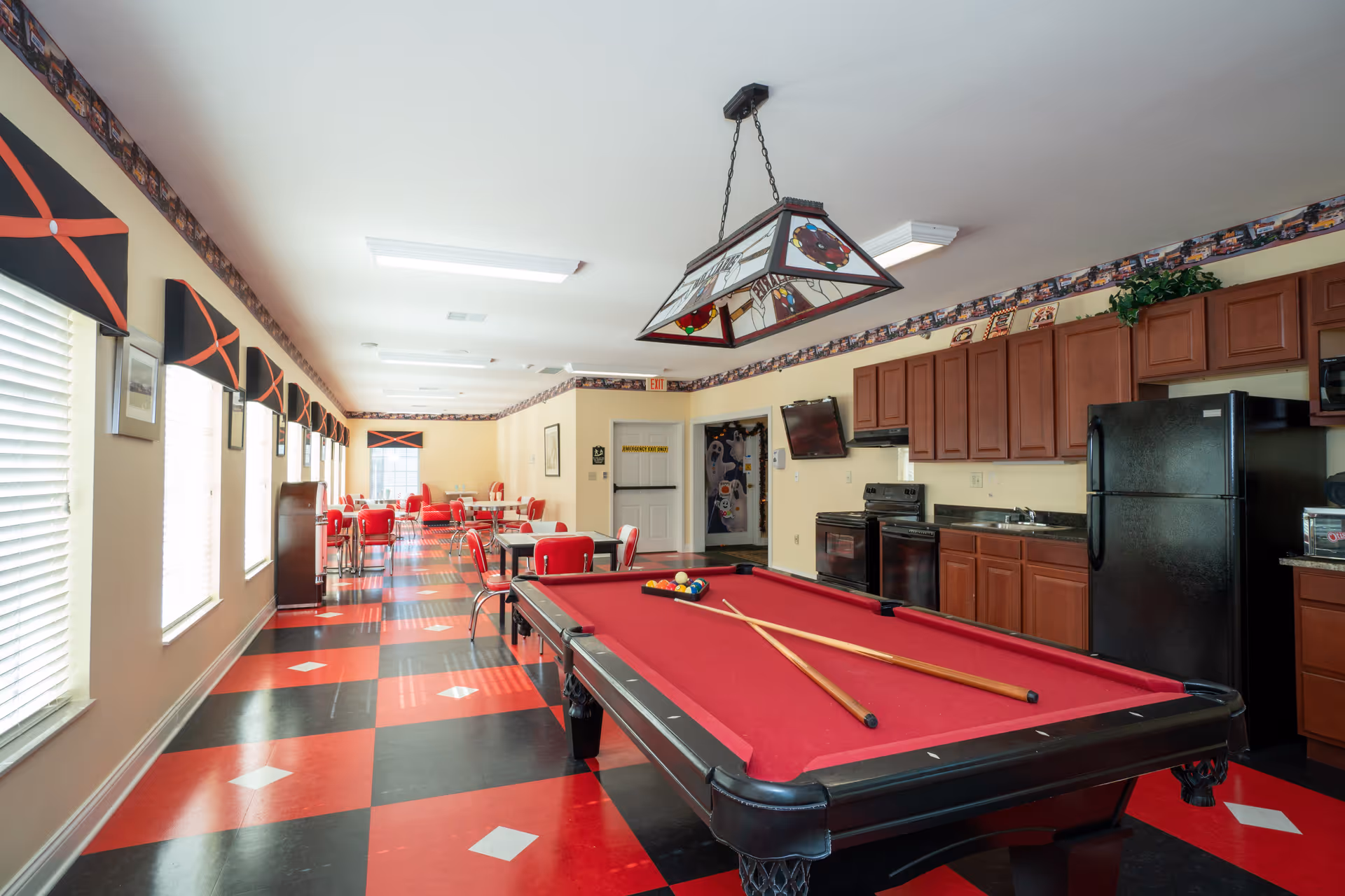Interior view of a recreational and dining area featuring a red pool table with two cues and balls, a kitchen area with wooden cabinets, black refrigerator, stove, and microwave. The room has a red and black checkered floor, multiple windows with black and red valances, several tables and chairs arranged for dining or socializing, and a hanging stained glass light fixture above the pool table.