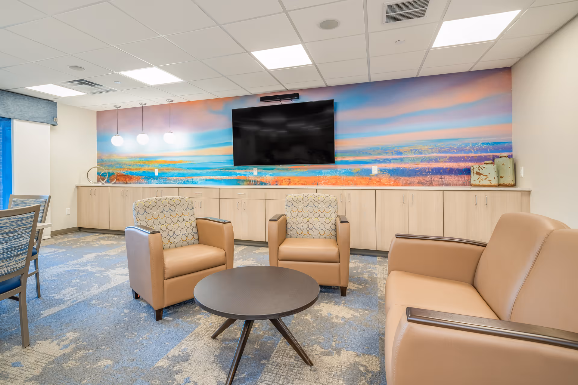 A modern lounge area with two patterned armchairs, a tan leather sofa, and a round dark wood coffee table. The back wall features a large colorful mural with shades of blue, orange, and pink, and a flat-screen TV mounted above light wood cabinets. The room has a blue and beige patterned carpet and a ceiling with recessed lighting and three hanging globe lights.