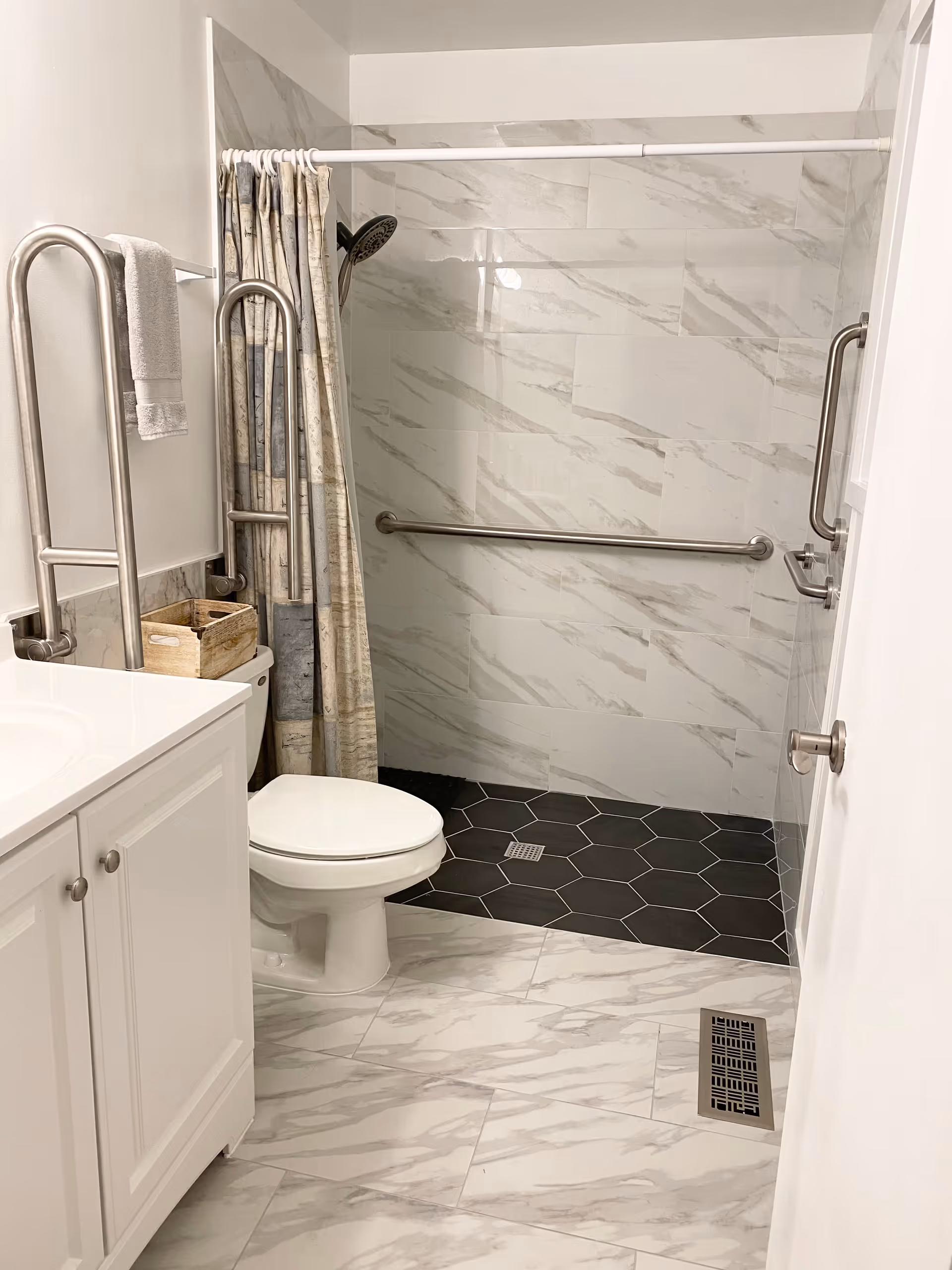 A clean and modern bathroom with white marble-patterned tiles on the floor and walls. There is a white toilet next to a white vanity with a sink. The shower area has dark hexagonal floor tiles and a wall-mounted showerhead with a curtain. Stainless steel grab bars are installed near the toilet and inside the shower for accessibility.