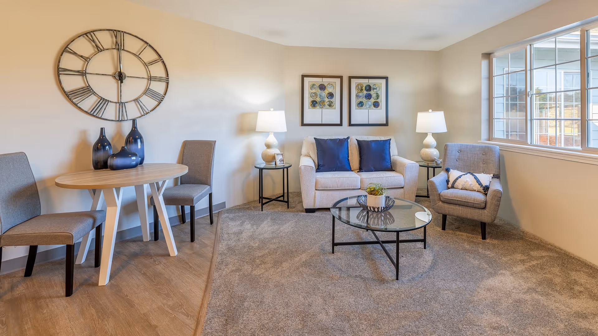 A cozy living room with a beige loveseat adorned with blue pillows, a gray armchair with a decorative pillow, and a round glass coffee table with a small plant. Two table lamps on either side of the loveseat provide warm lighting. On the left side, there is a small wooden table with two gray chairs and three decorative blue vases. A large wall clock and two framed artworks hang on the beige walls. A large window lets in natural light, illuminating the carpeted floor and wooden flooring near the table.