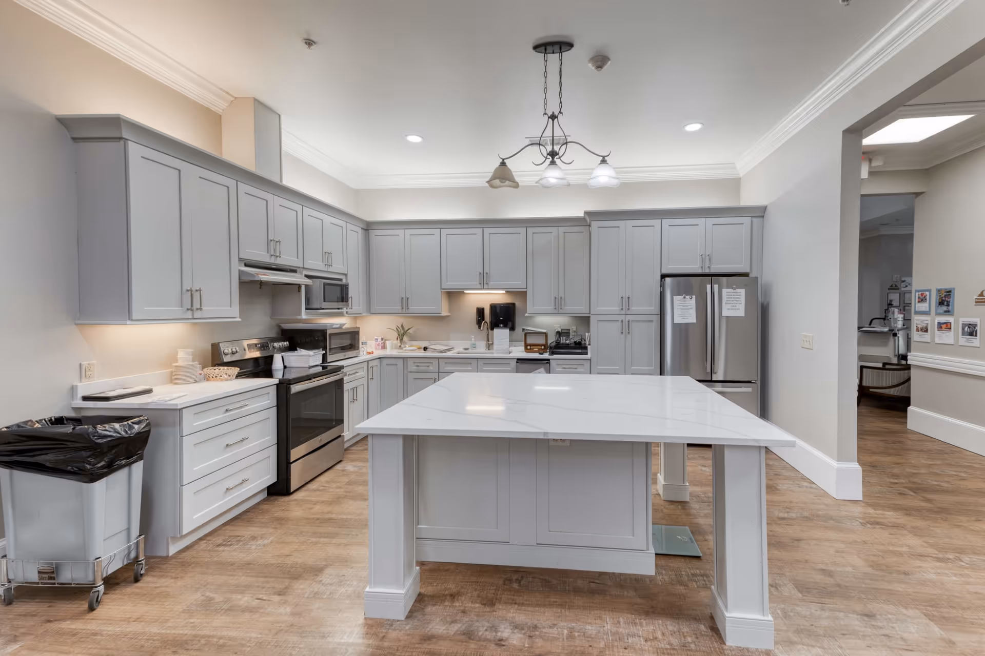 A modern kitchen with light gray cabinets, a large white island countertop in the center, stainless steel appliances including a refrigerator, stove, and microwave. The kitchen has wood flooring and is well-lit with ceiling lights and under-cabinet lighting. There is a trash bin on the left side and an open doorway on the right leading to another room.